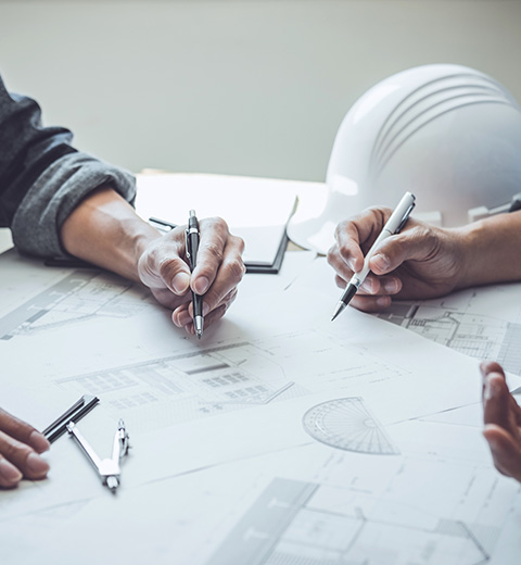 hands of three people holding pens discussing building plans with a construction helmet on the table